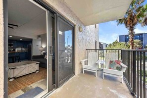 A balcony with a sliding glass door leading to a modern kitchen, a white chair with a floral cushion, and a palm tree in the background.