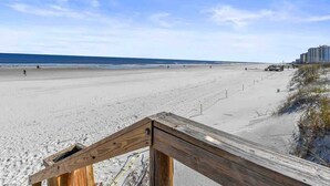 A beach with a wooden walkway leading to the water, a clear sky, and a distant building.