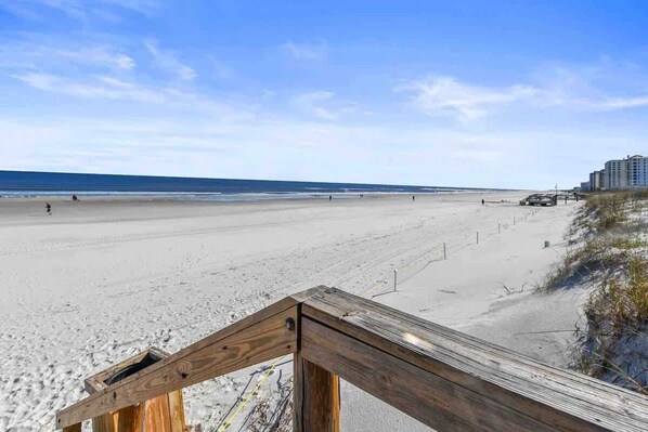 A beach with a wooden walkway leading to the water, a clear sky, and a distant building.