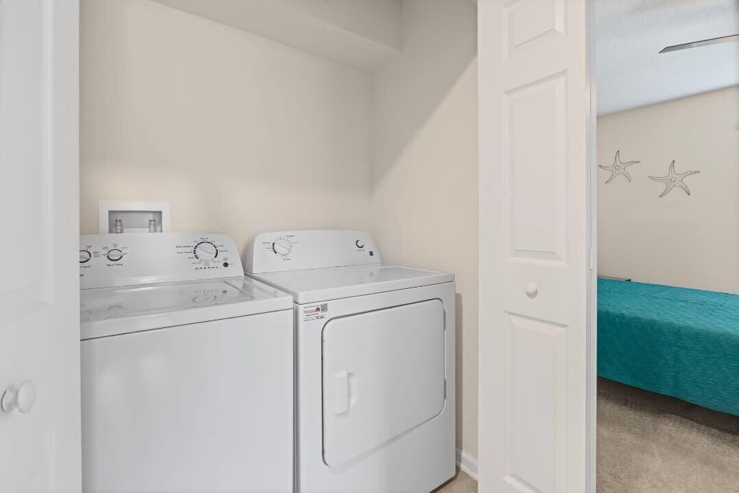 A laundry room with two washing machines and a dryer, a teal bed in the adjacent room, and a white door.