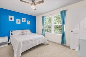 Ein Schlafzimmer mit Bett, Deckenventilator, Fenster mit Vorhängen und Wandbildern.