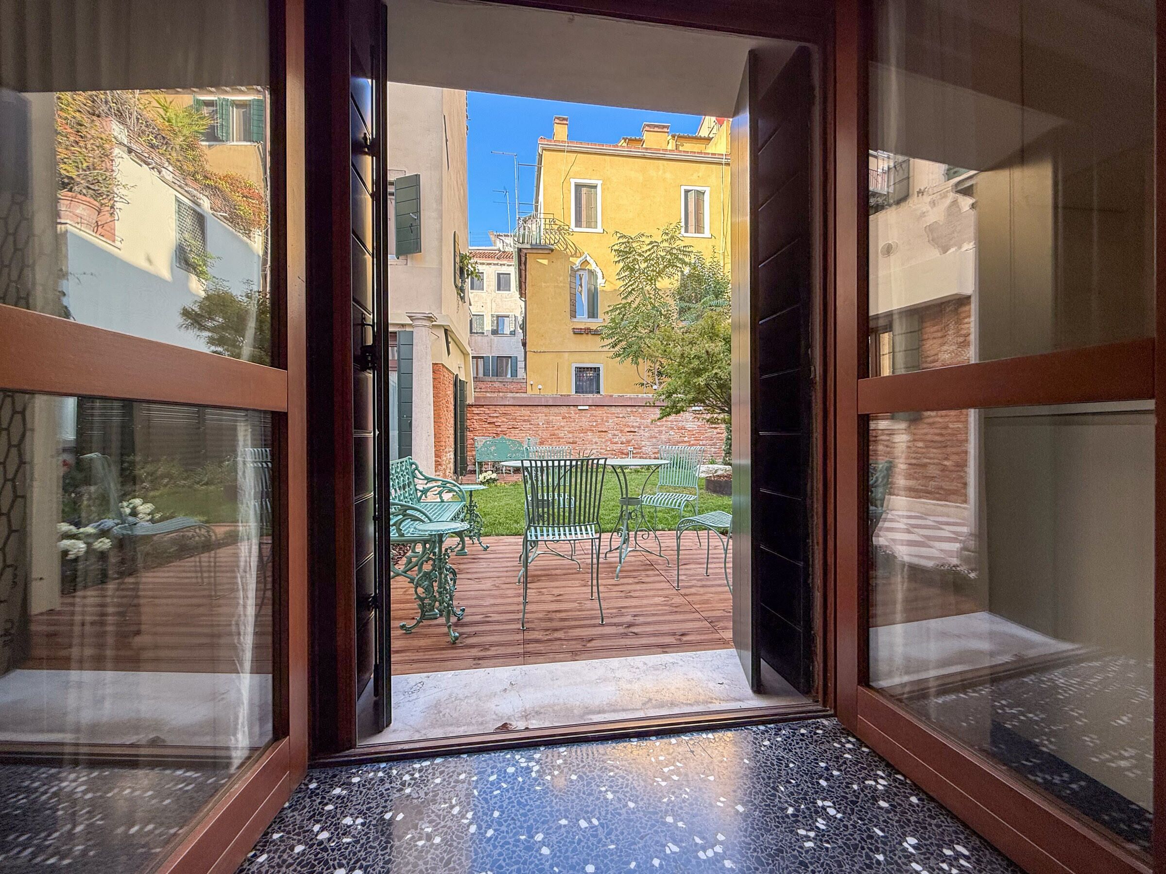 An open door leading to a courtyard with a tiled floor, outdoor furniture, and a view of buildings.