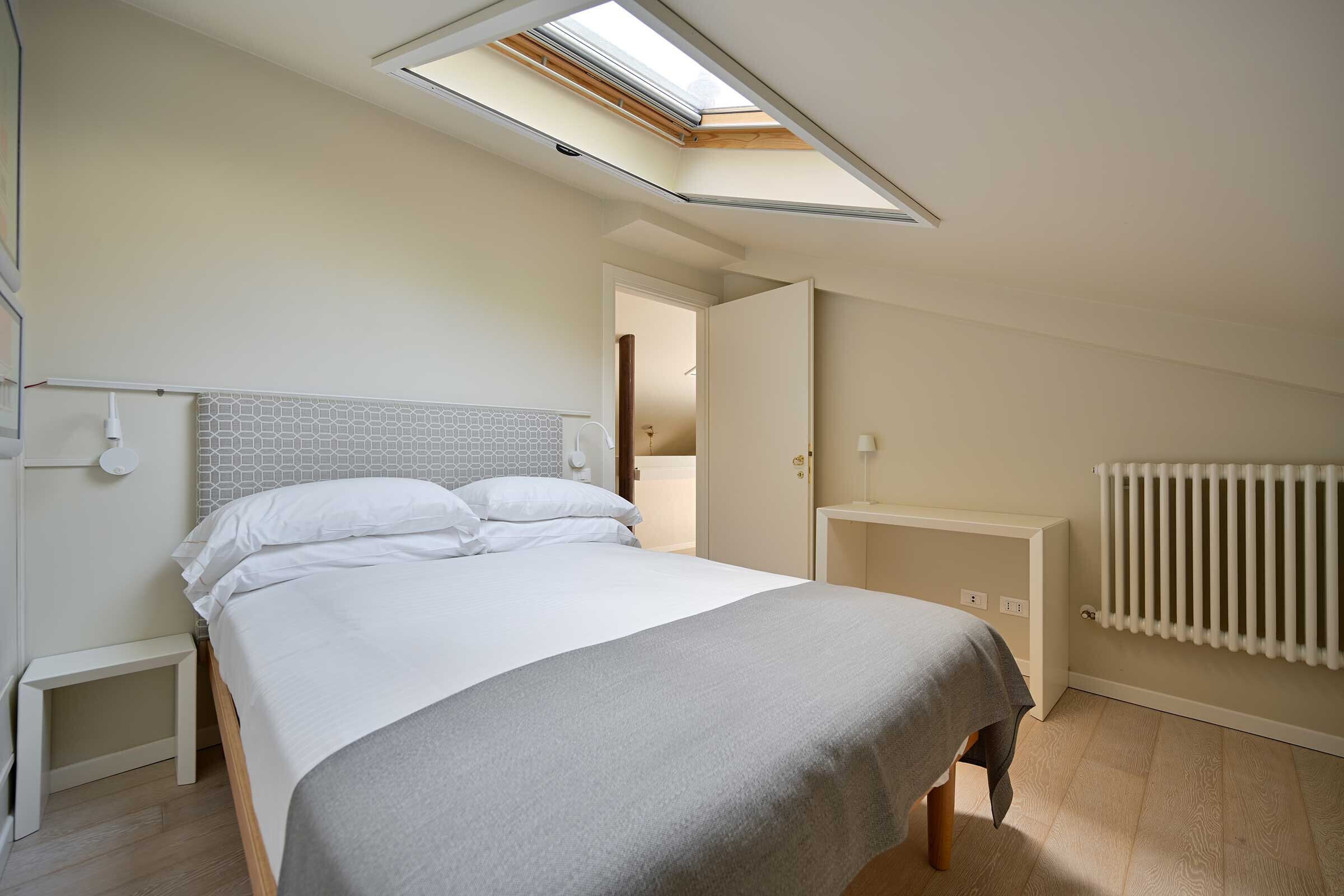 Une chambre avec une lucarne, un lit avec une literie blanche, une table de chevet, un radiateur et un mur recouvert de carreaux.