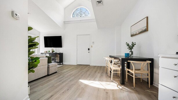 Una habitación amplia con techo alto, un comedor con mesa y sillas, un sofá, un televisor y una puerta.