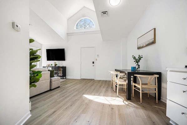 Ein geräumiger Raum mit hohen Decken, einem Essbereich mit Tisch und Stühlen, einer Couch, einem Fernseher und einer Tür.