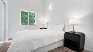 Un dormitorio con una cama, dos mesitas de noche, un ventanal y una lámpara fijada a la pared.