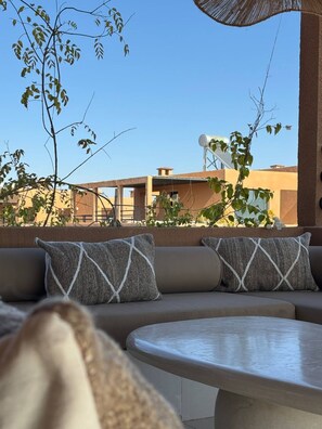 An outdoor seating area with a sofa and a round table, surrounded by plants and a clear view of buildings in the background. - Villa Camelia - Breakfast and Heated Pool (Marrakech)
