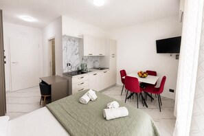 Ein kompaktes Hotelzimmer mit Bett, kleinem Schreibtisch, einer Küchenzeile, einem Essbereich mit roten Stühlen und einem an der Wand befestigten Flachbildfernseher.
