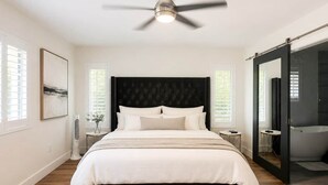 Et soveværelse med seng, loftventilator, natbord, ventilator på væggen og en skydedør til et badeværelse.