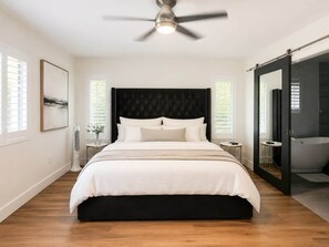 Ein Schlafzimmer mit Bett, Deckenventilator, Nachttisch, Wandventilator und einer Schiebetür zum Badezimmer.
