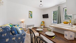 Una stanza moderna e luminosa con un tavolo da pranzo apparecchiato per due, un divano, una TV a parete e una finestra con vista sul verde.