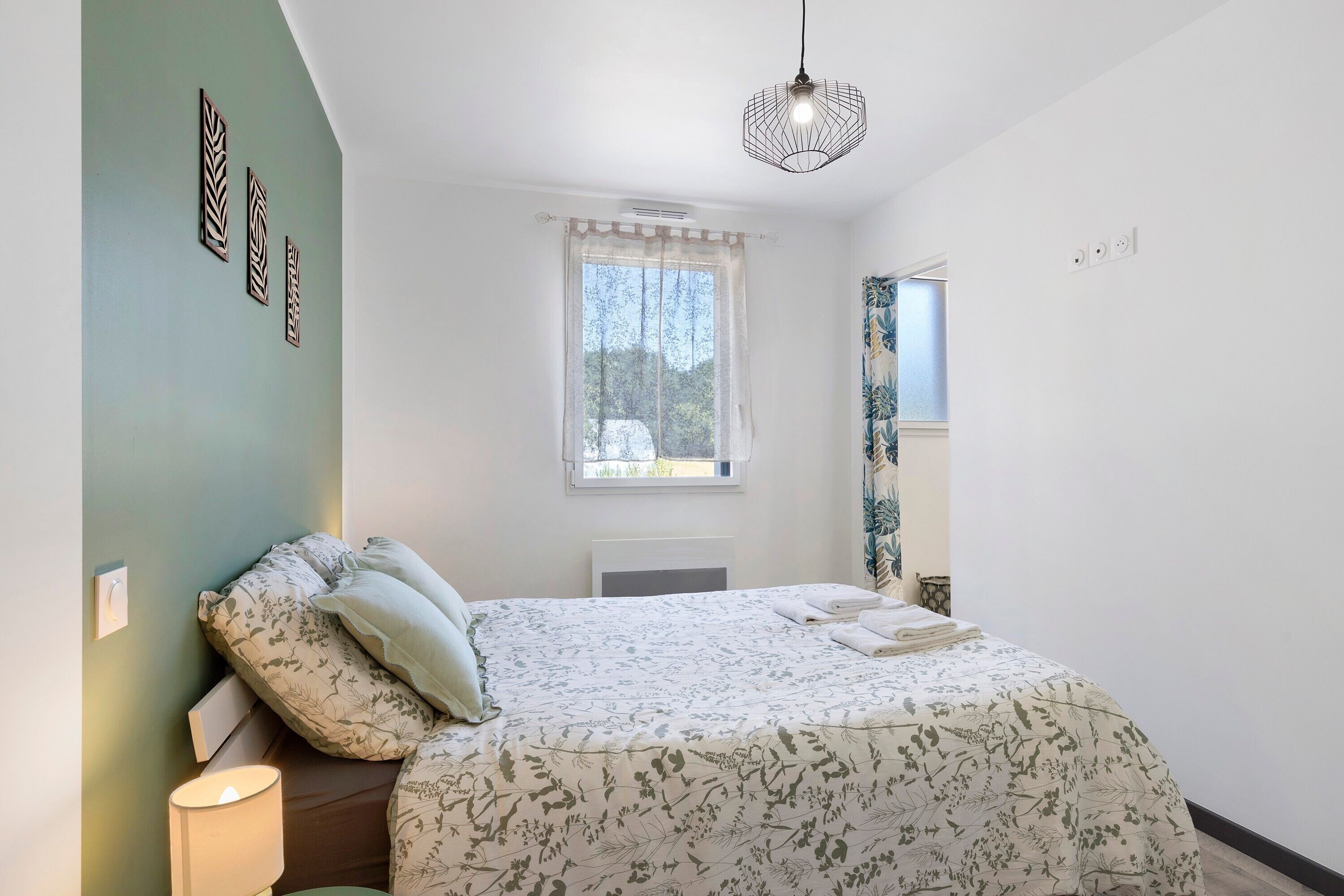 A bedroom with a bed, a bedside lamp, a window with a view, and a hanging light fixture.