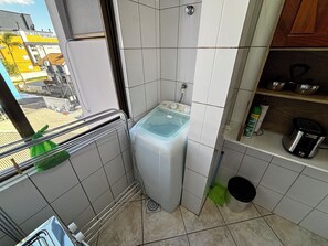 A compact bathroom with a washing machine, a small shelf with kitchen items, and a view of the outside. - Unleash your best self by the sea! (Florianópolis)