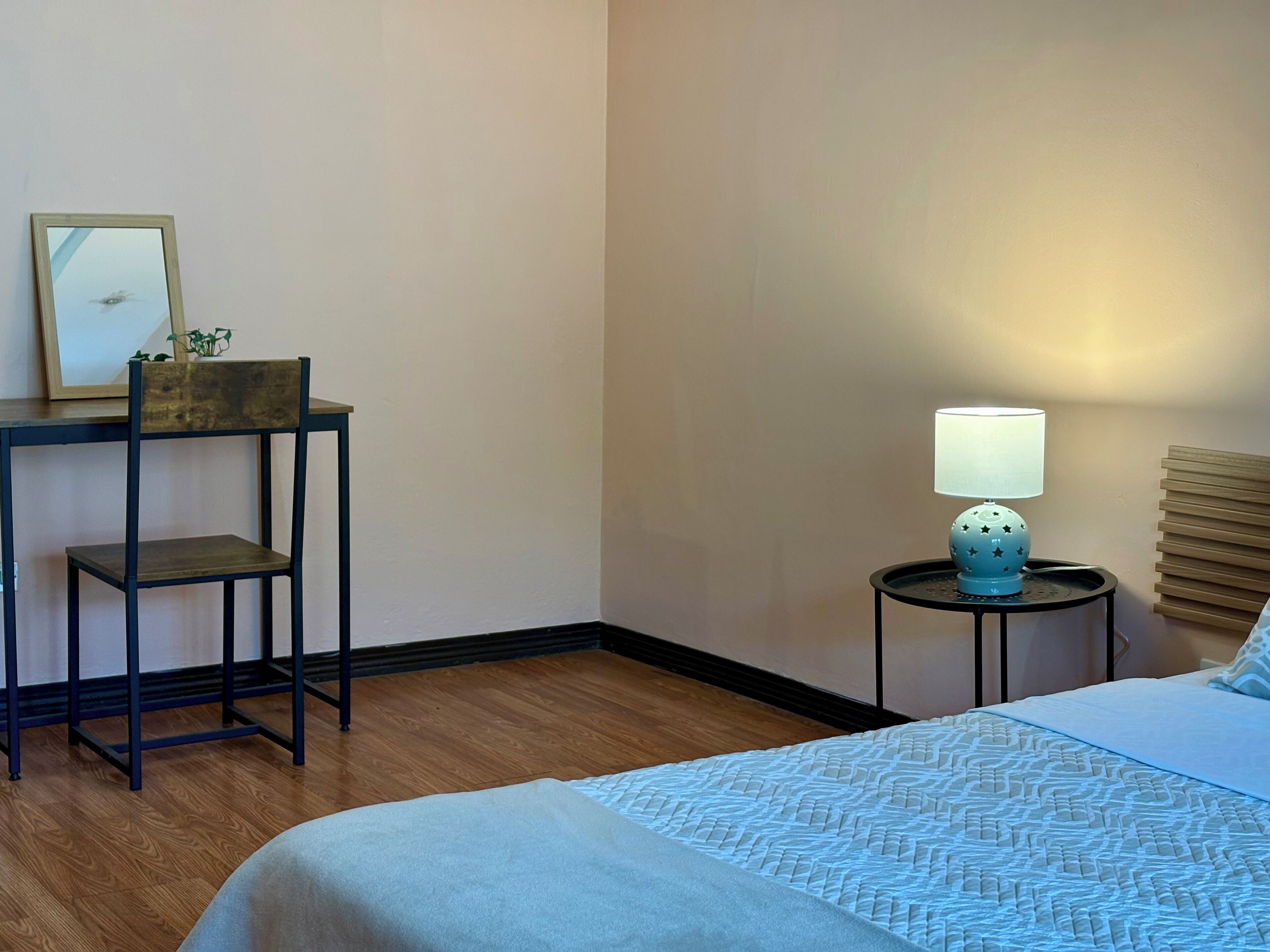 A hotel room with a bed, a desk with a chair, a lamp, and a picture frame on the wall.