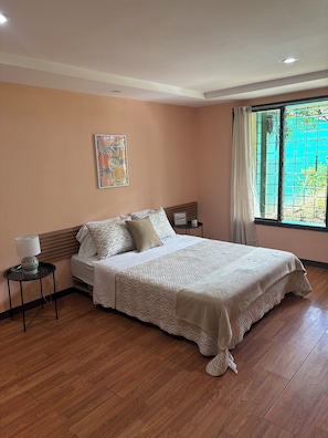A bedroom with a bed, a bedside table with a lamp, a window with a view, and a wall with a framed picture.