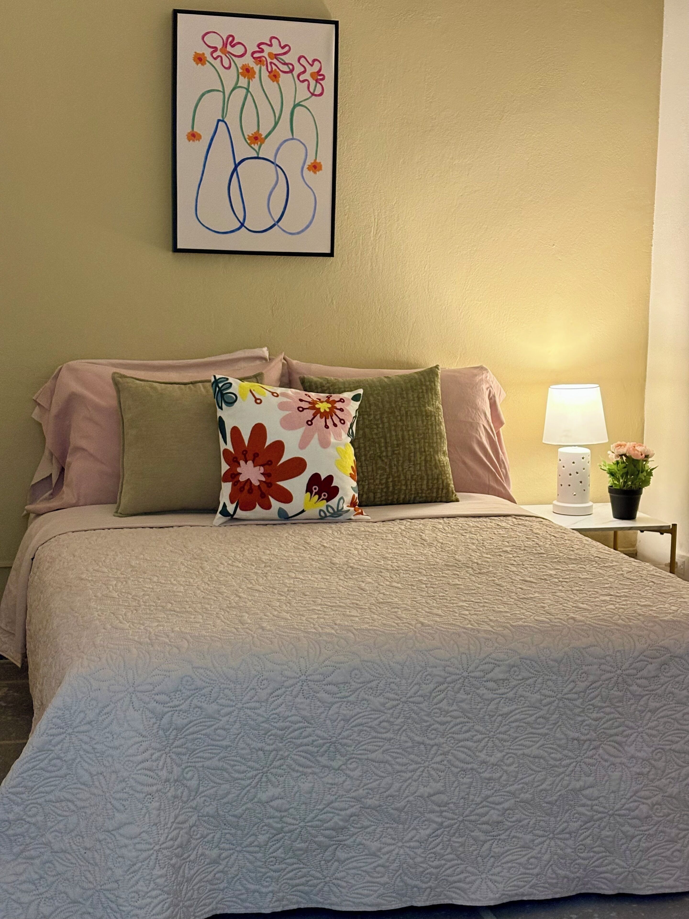 Una cama bien hecha con almohadas estampadas con motivos florales, una obra de arte colgada en la pared, una lámpara de noche y una pequeña planta en maceta.
