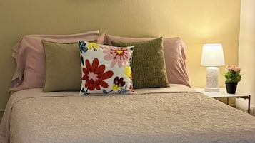 Una cama bien hecha con almohadas estampadas con motivos florales, una obra de arte colgada en la pared, una lámpara de noche y una pequeña planta en maceta.