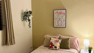 Un dormitorio con cama, almohadas, una mesita de noche con lámpara, una planta en la pared y una obra de arte enmarcada.