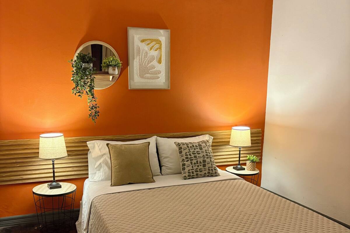 A bedroom with an orange wall, a bed with a beige quilt, two bedside lamps, a round mirror, and a framed artwork.