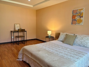 A bedroom with a bed, a desk, a chair, a nightstand with a lamp, and a wall art piece.