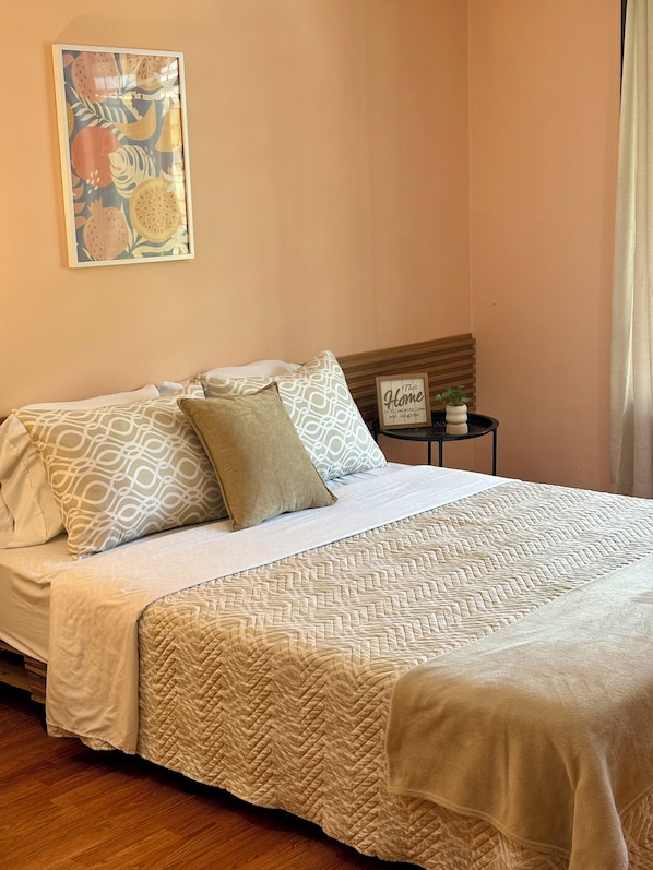 A neatly made bed with patterned pillows and a quilted bedspread, a small bedside table with a plant, and a wall art piece.