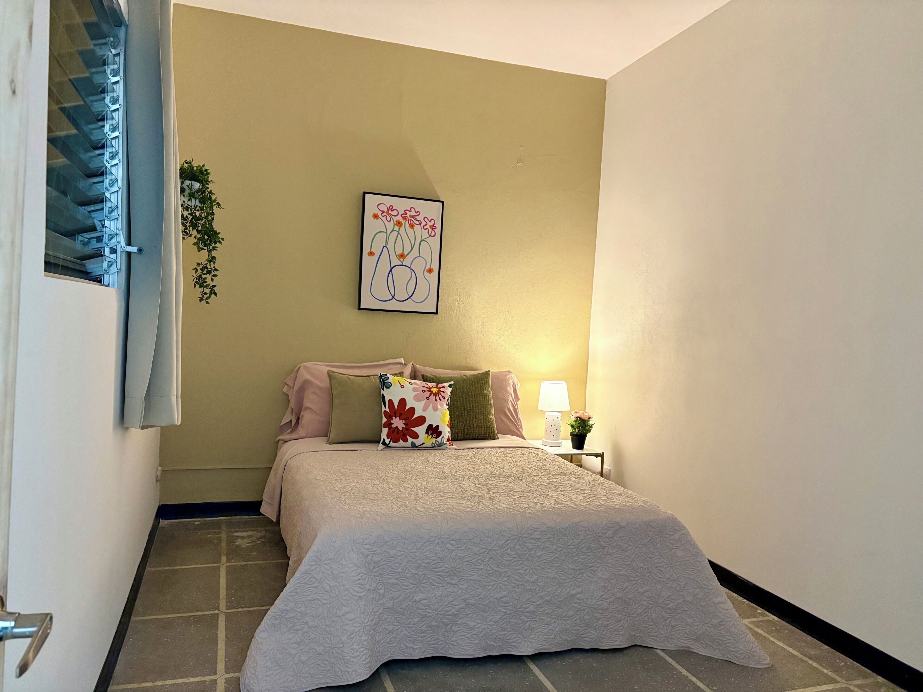 A bedroom with a bed, pillows, a wall-mounted artwork, a nightstand with a lamp, and a potted plant.