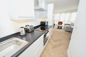 A modern kitchen with a black countertop, stainless steel sink, and white cabinetry. There is a dining area with a grey sofa and a small round table. - Modern & Bright 1BED Apartment in Leeds - Sleeps 3 (Leeds)