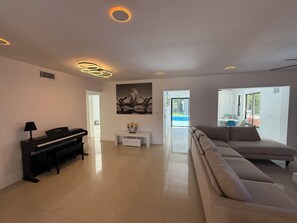 A modern living room with a piano, a sofa, a dining area, and a view of the outdoors. - Beautiful Oasis Private Pool (Plantation)