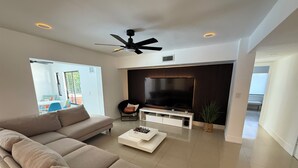 Una sala de estar moderna con un sofá seccional, un televisor de pantalla plana, un ventilador de techo y un ventanal grande.