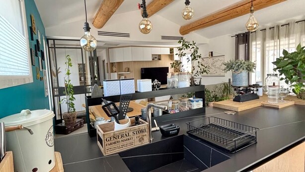Ett modernt kök med en svart bänkskiva, träbjälkar, hängande lampor och utsikt över matplatsen.