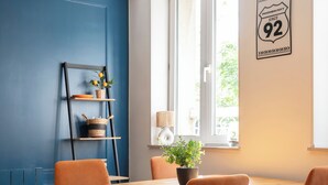 A dining area with a wooden table and orange chairs, a blue wall, a window with a view, and a wall-mounted sign. - Rock in share - Le Graoully (Metz)