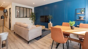 A modern living room with a sofa, dining table, chairs, a TV, and a blue accent wall. - Rock in share - Le Graoully (Metz)