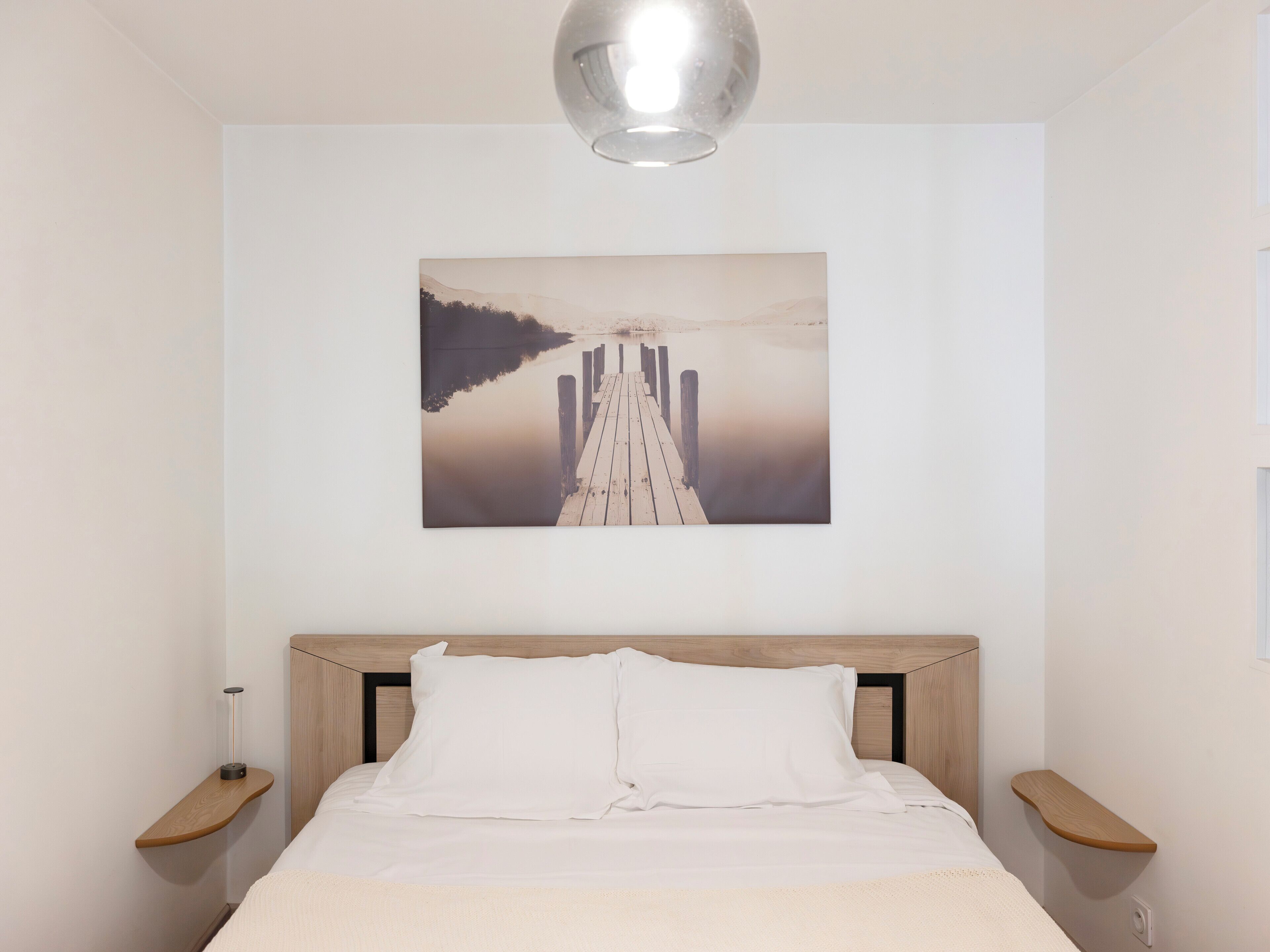 A bedroom with a wooden headboard, a white bedspread, and a painting of a pier extending into a calm body of water.