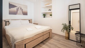 A modern bedroom with a wooden bed, a bedside table, a hanging light, a wall-mounted shelf with plants, and a door with a mirror. - Rock in share - Le Graoully (Metz)