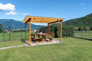 Una pèrgola de fusta amb una taula i bancs en una zona verda, amb muntanyes al fons.