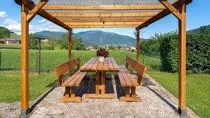 A wooden pergola with a table and benches, set on a stone patio with a view of mountains.