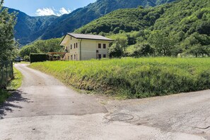 A rural road leading to a two-story building with a sloped roof, surrounded by lush greenery and mountains. - Room 'Monte Grappa View' with Mountain View, Wi-Fi and Air Conditioning (Alano di Piave)