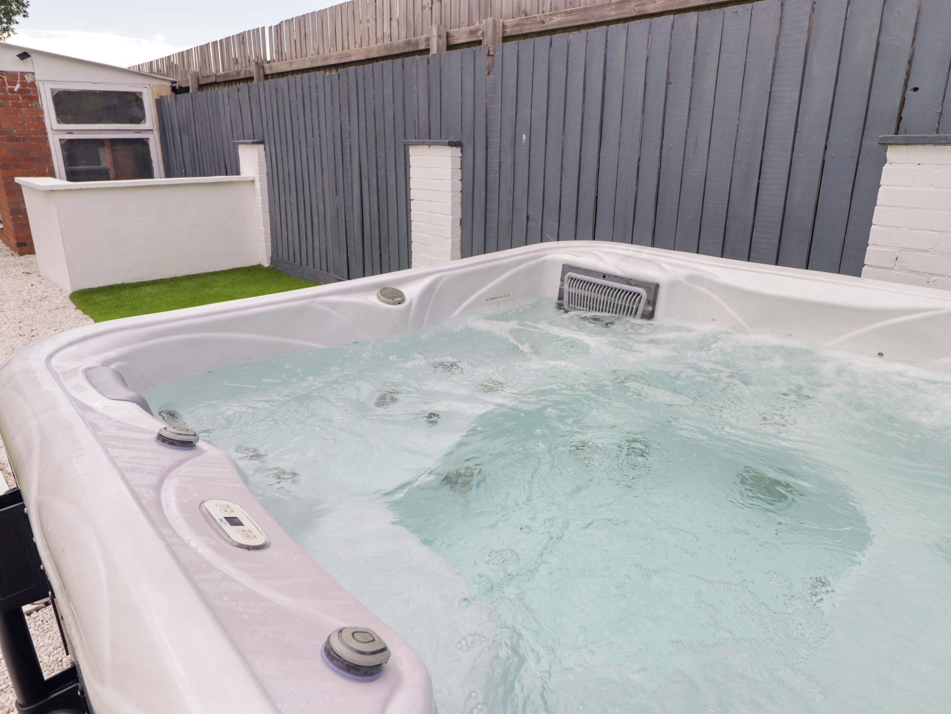 A hot tub with a control panel, surrounded by a white wall and a grey fence.