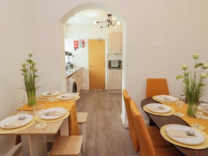 Un comedor con una mesa larga dispuesta para una comida, una cocina al fondo y una puerta que da a otra habitación.