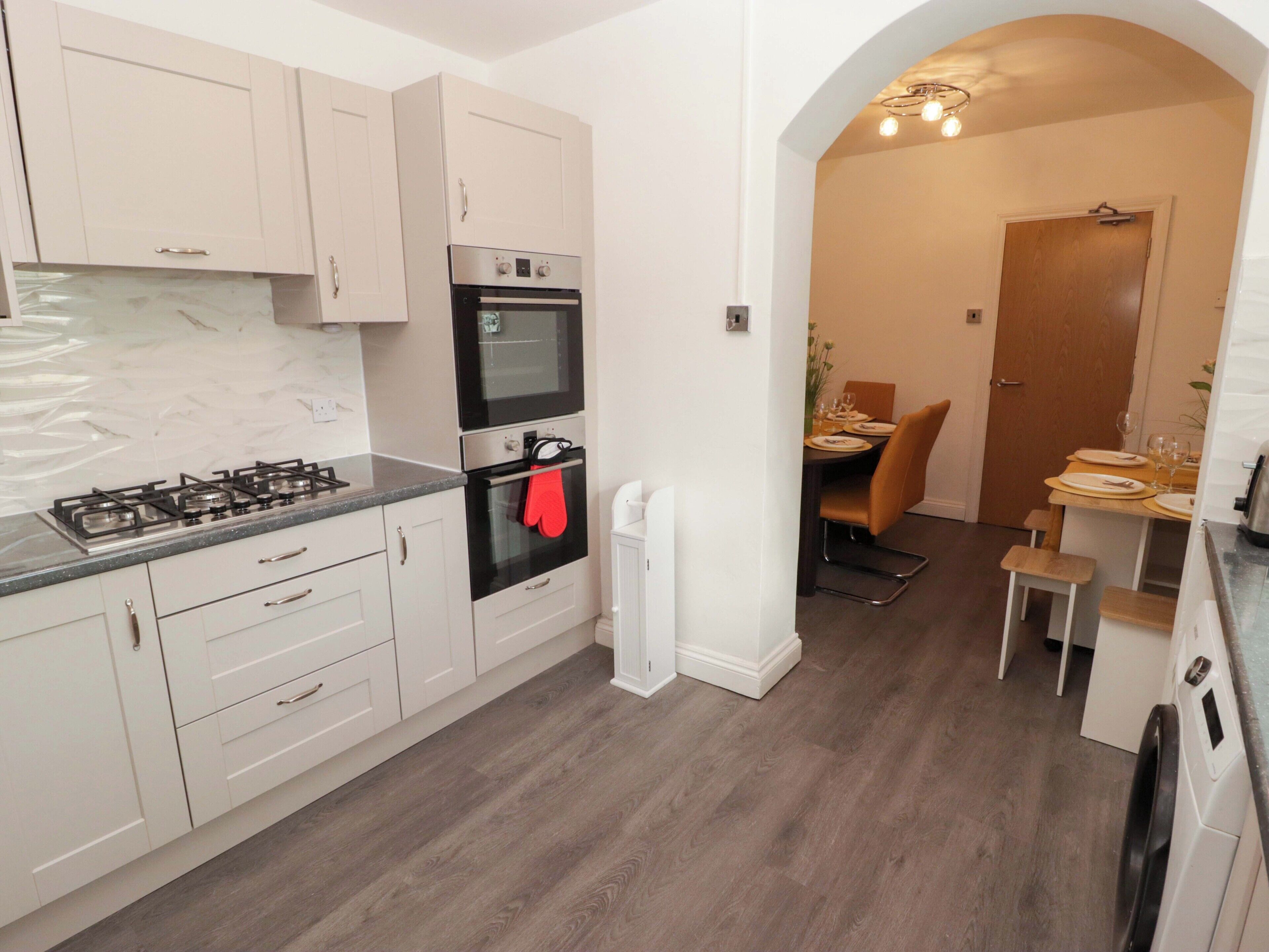 Een moderne keuken met witte kastjes, een gasfornuis, een oven en een eethoek met een houten tafel en stoelen.