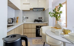 A compact kitchen with a coffee maker, kettle, and a small oven. There is a dining area with a table set for two, and a potted plant on the window sill.