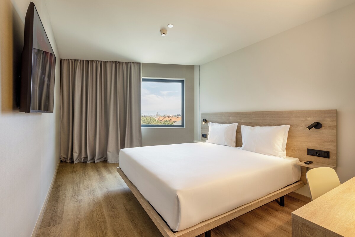 A hotel room with a large bed, a desk, a TV mounted on the wall, and a window with a view of the cityscape.
