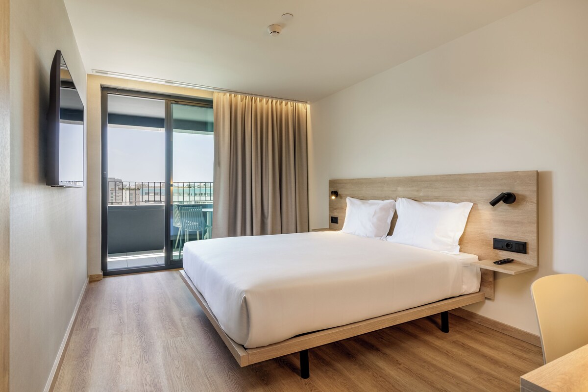 A hotel room with a large bed, a wooden headboard, a flat-screen TV mounted on the wall, and a sliding glass door leading to an outdoor area.