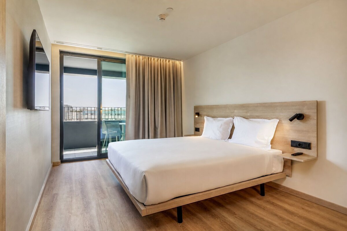 A hotel room with a large bed, a TV mounted on the wall, a balcony with a view, and a wooden floor.