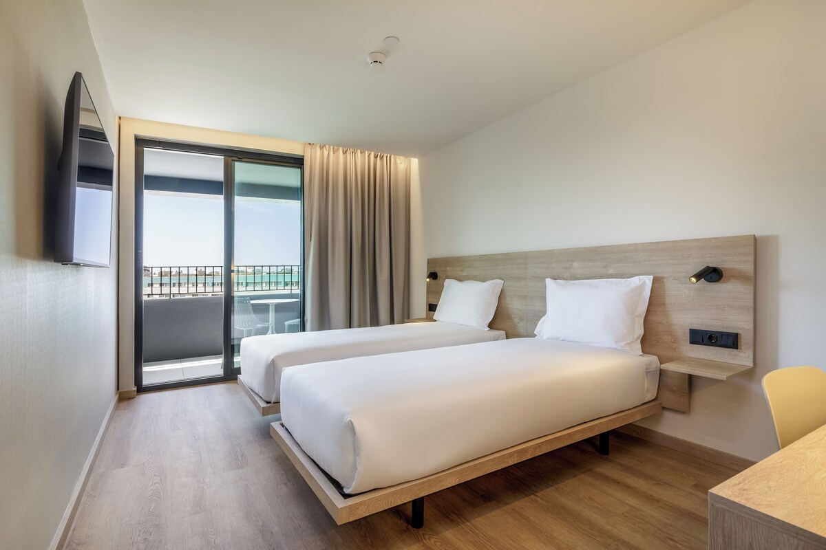 A hotel room with two beds, a TV, a desk, and a balcony with a view.