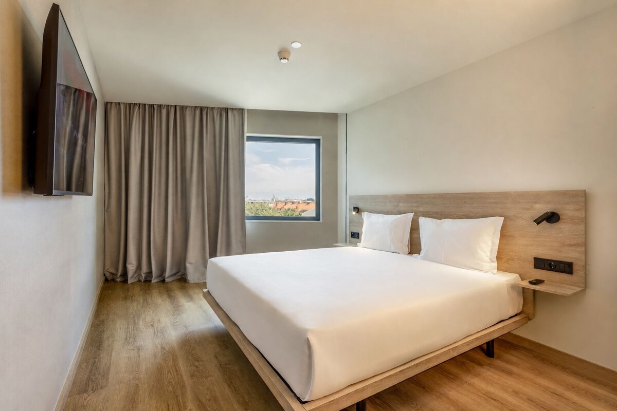A hotel room with a large bed, a mounted TV, a window with curtains, and a view of buildings outside.