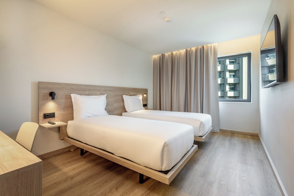 A hotel room with two beds, a desk, a chair, a TV mounted on the wall, and a window with curtains.