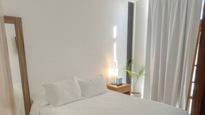 Ein ordentlich bezogenes Bett mit weißer Decke, ein Nachttisch mit Lampe, eine Zimmerpflanze und ein Spiegel an der Wand.
