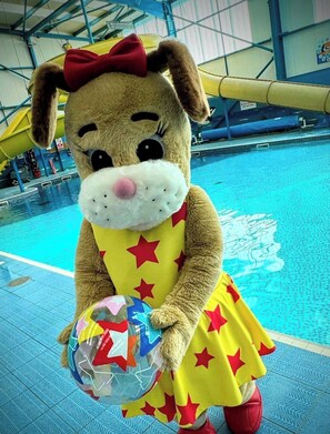 A mascot in a yellow dress with red stars, holding a colorful ball, standing by a pool.