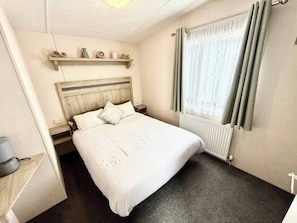 Habitación de hotel ordenada con cama, mesita de noche, estante con adornos, ventana con cortinas y radiador.